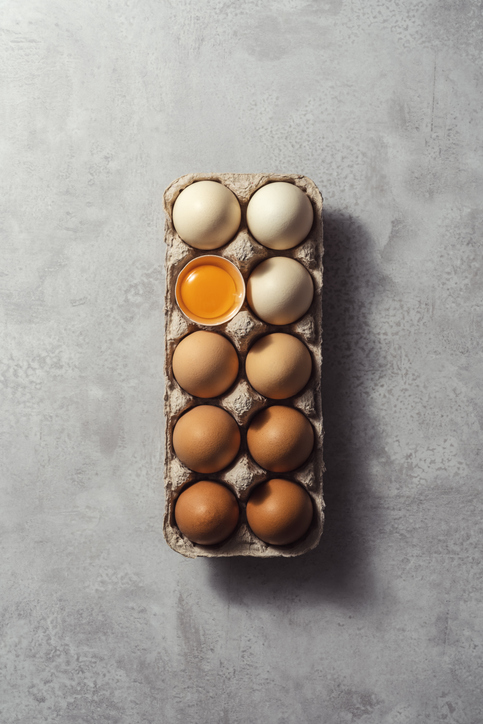 Box of color gradient brown eggs with one cracked egg, rectangular egg carton on grey background. Easter eggs. Shot from directly above.